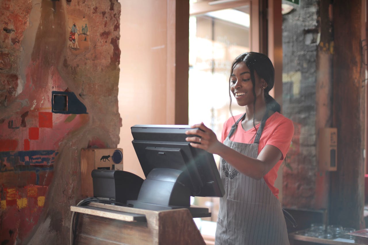 team-03 Cheerful American African waitress in apron working on counter monitor while registering order at cozy cafe